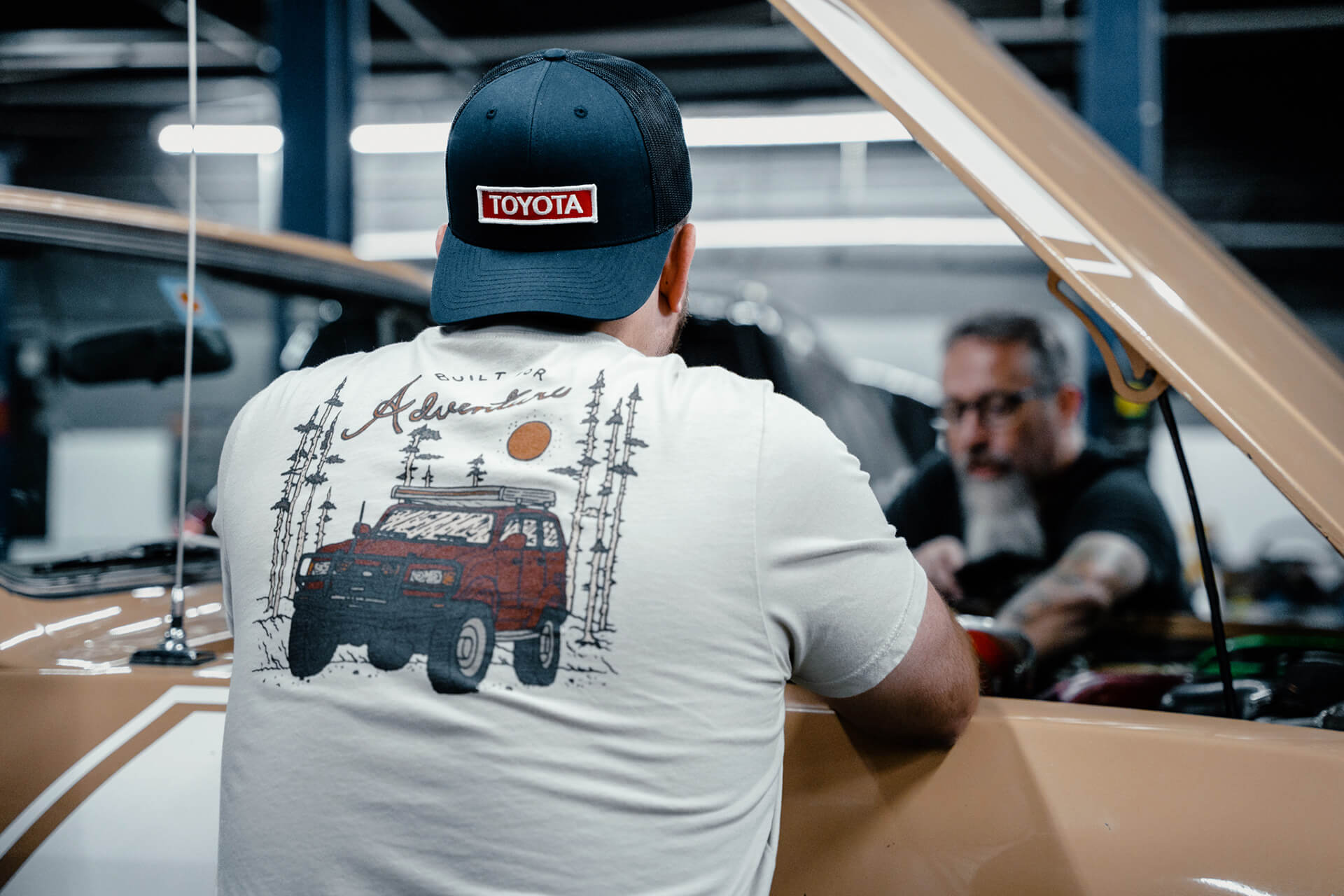 Picture from behind showing the back of a white built for adventure shirt and a blue trucker hat with a red and white Toyota patch